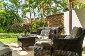 A wicker outdoor seating area with a sofa and ottoman, a small table with a vase and flowers, and a view of a lush garden with palm trees and tropical plants. - Garden oasis, spacious lanai & open concept living (Kihei)