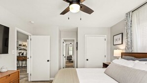 Un dormitorio con cama, ventilador de techo, mesita de noche con lámpara y vistas a un pasillo que lleva a un baño.
