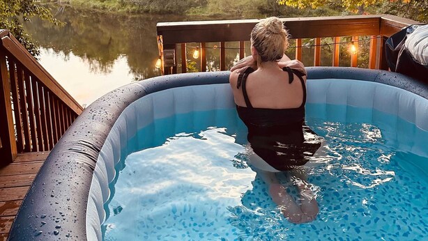 Eine Person sitzt in einem aufblasbaren Pool mit einer Holzterrasse und Blick auf einen von Bäumen umgebenen See.
