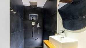 A modern bathroom with dark tiles, a wooden countertop, a white sink, a mirror, and a shower with a handheld showerhead. - 1 Bedroom Apartment in Hattingen (Hattingen)