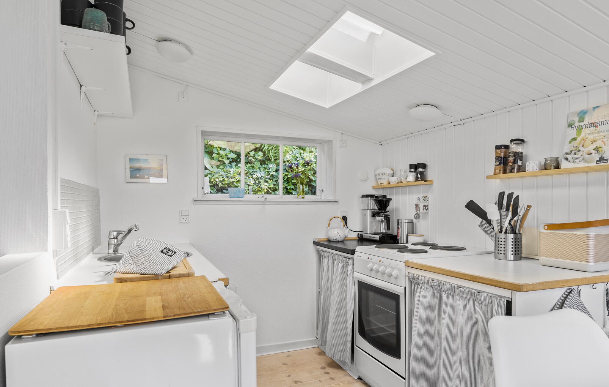Une cuisine compacte avec des meubles blancs, une fenêtre donnant sur un espace verdoyant, une machine à café et une planche à découper en bois posée sur le plan de travail.
