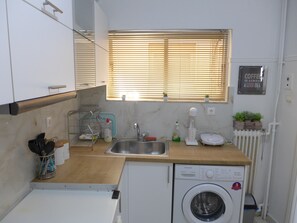 A compact kitchen with a washing machine, a sink, and a window with blinds. - Koum Kapi 1st floor apt (Chania)