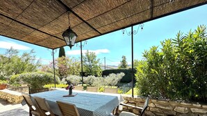 Eine Terrasse im Freien mit Tisch und Stühlen, einem hängenden Laternenlicht und Blick auf einen Garten mit verschiedenen Pflanzen und einer Hecke.