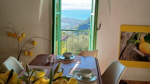 A dining area with a table set for two, a view of the sea through an open window, and a vase of yellow flowers.