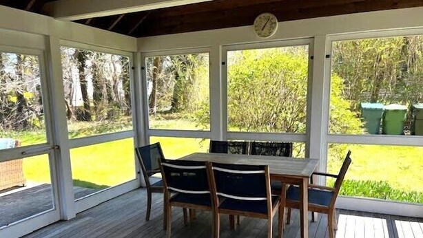 Une terrasse couverte avec une table et des chaises en bois, de grandes fenêtres et une horloge au mur.