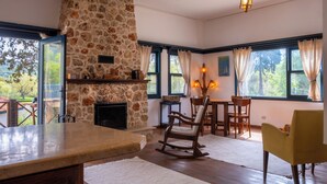 Camino in pietra, tavolo da pranzo in legno e sedie in una stanza con ampie finestre e vista sugli alberi.