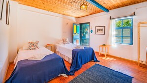 Una stanza con due letti, travi in legno a soffitto, una finestra con persiane blu e una porta con pannelli di vetro colorato.