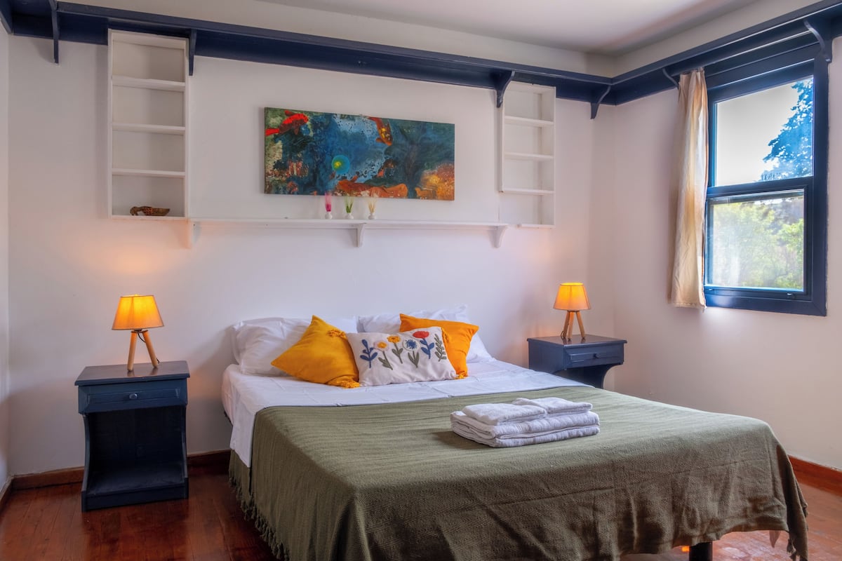 Une chambre à coucher bien rangée, avec un lit, deux tables de chevet dotées de lampes, une étagère ornée d’objets décoratifs, un tableau et une fenêtre avec des rideaux.