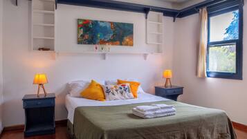 A neatly arranged bedroom with a bed, two bedside tables with lamps, a shelf with decorative items, a painting, and a window with curtains.