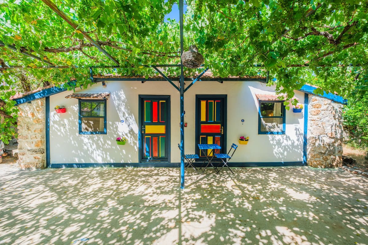 Une petite maison avec un toit en tuiles, deux portes aux encadrements colorés et une petite table avec des chaises à l’extérieur.