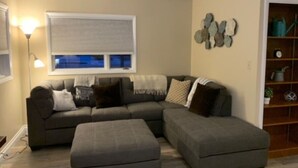 A living room with a sectional sofa, an ottoman, a wall-mounted shelf with decorative items, and a window with blinds. - Longhorn Lodge - Extended Stay (Great Falls)