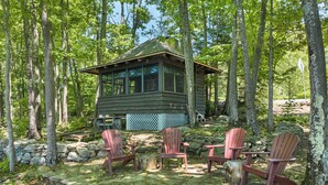 A cabin with a stone wall and wooden deck, surrounded by trees. - 5 Bedroom Squam Lake Home with Dock Paddleboard Kayak and canoe Sleeps 17 (Center Sandwich)