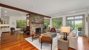 A spacious living room with a stone fireplace, wooden beams, a sofa, a coffee table, and a view of the outdoors through large windows. - 5 Bedroom Squam Lake Home with Dock Paddleboard Kayak and canoe Sleeps 17 (Center Sandwich)