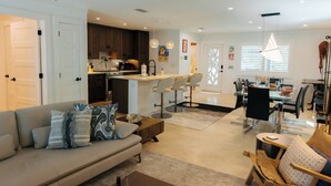 A modern living room with a sofa, a dining area with a glass table, and a kitchen with dark cabinets and pendant lighting. - Grayt Balls of Fire 4 bed, 4 1/2 bath, private pool  Grayton Beach (Santa Rosa Beach)