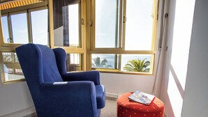 A blue armchair and a small round table with a red floral tablecloth in a room with large windows offering a view of palm trees and the sea. - First line apartment with sea view (Lo Pagán)