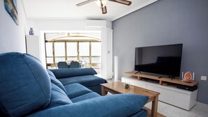 A modern living room with a blue sofa, a wooden coffee table, a flat-screen TV on a white cabinet, and a balcony with a railing. - First line apartment with sea view (Lo Pagán)