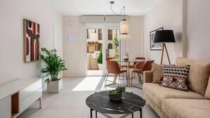 Un salon moderne avec un canapé, une table basse et un coin repas avec des chaises. On aperçoit une plante sur le côté et une lampe sur la droite.