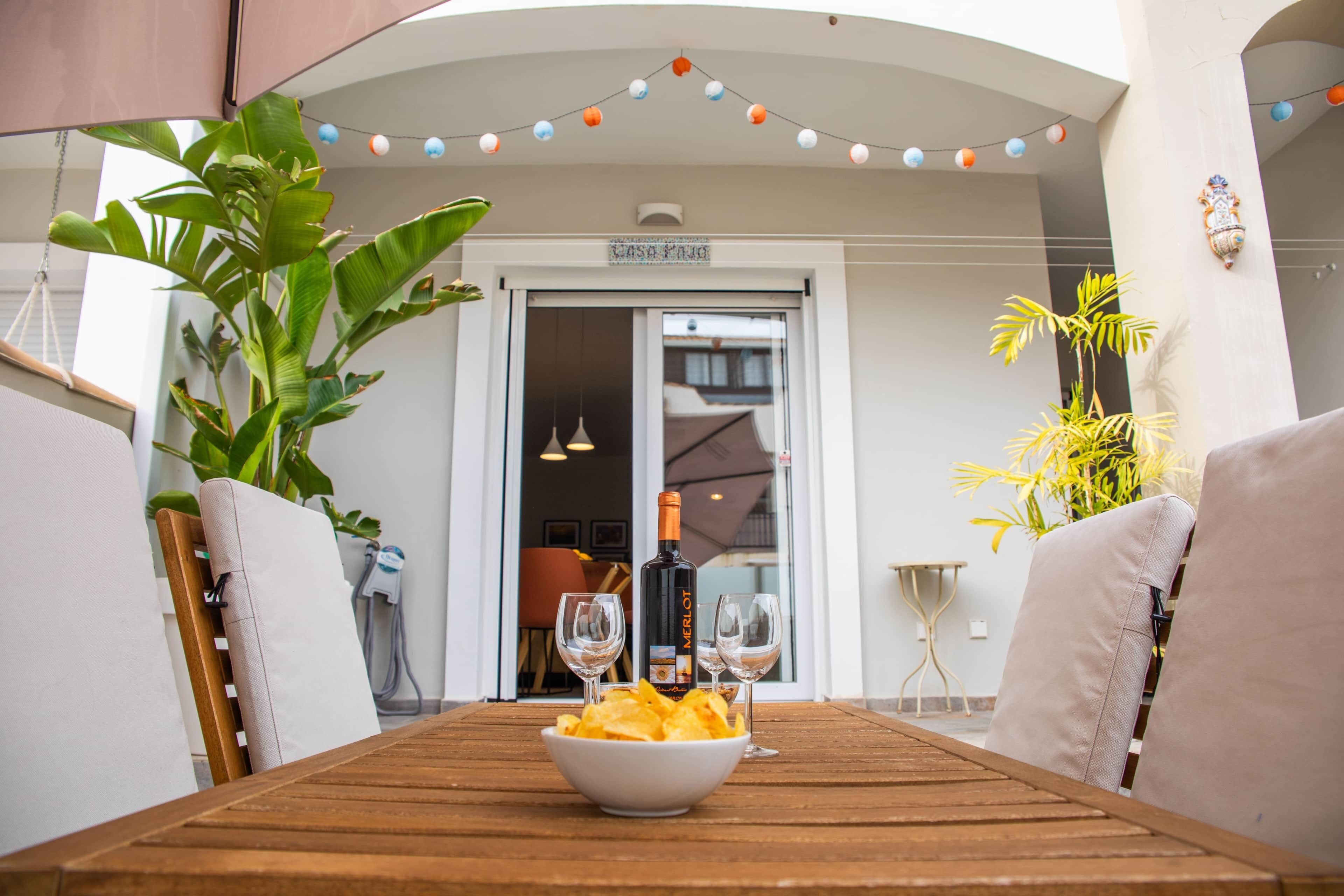 A wooden table with a bowl of chips and two wine glasses, a bottle of Merlot, and a string of colorful ornaments.