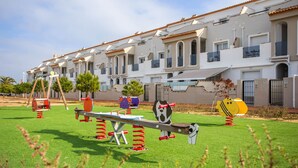 Un terrain de jeux avec des balançoires et une bascule, situé devant une rangée d’immeubles blancs dotés de balcons.