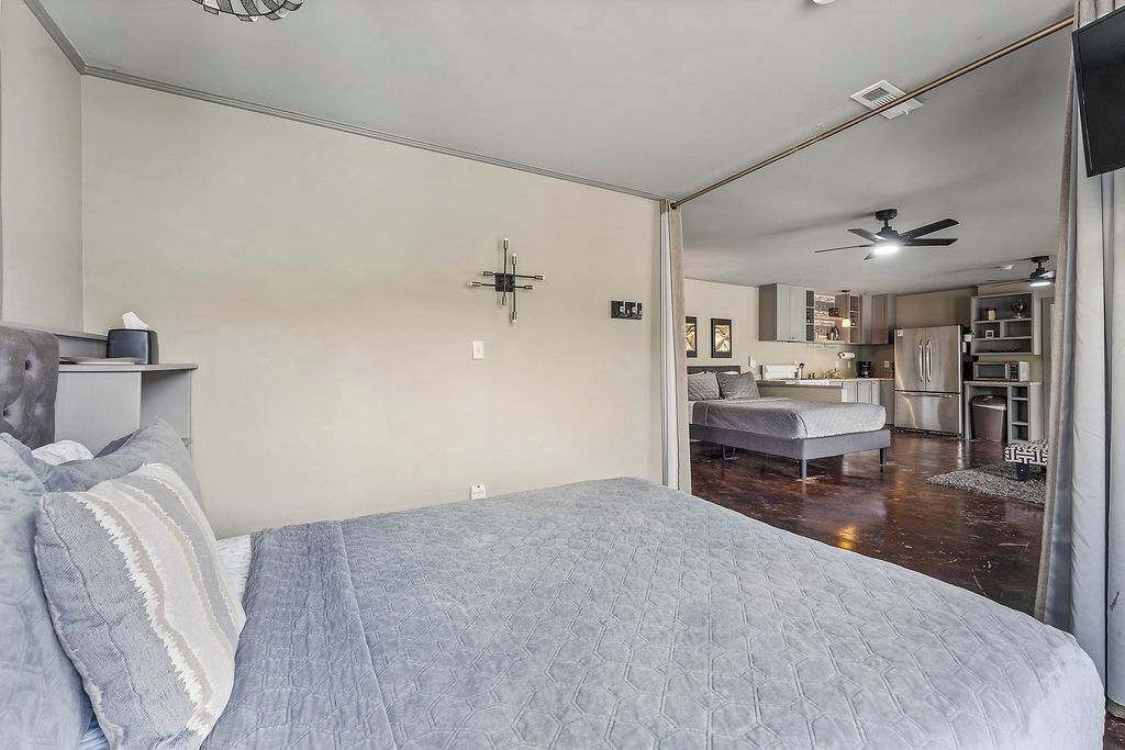 Une chambre avec un grand lit, une table de chevet, une lampe fixée au mur et une vue sur la cuisine.