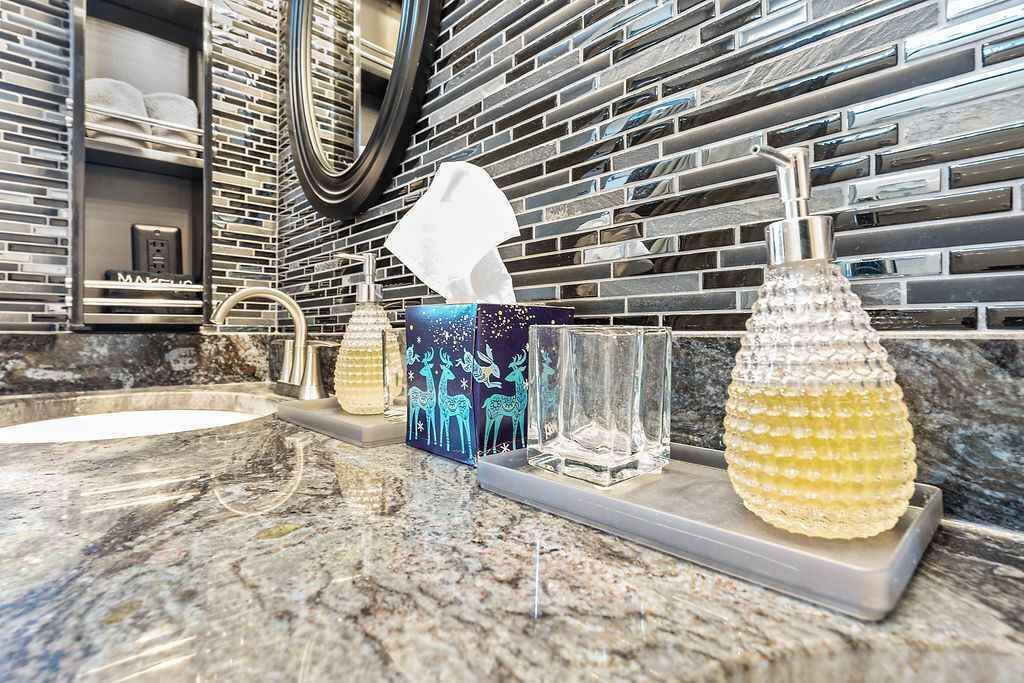 A bathroom with a modern design, featuring a granite countertop, a glass-topped sink, a mirror, and a decorative wall with a mosaic pattern.