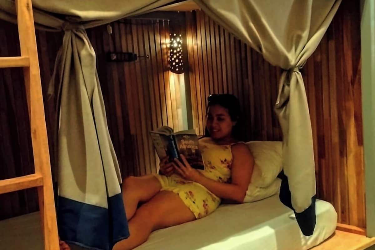 Uma pessoa lendo um livro em um quarto de madeira com beliche e uma gaveta pequena.