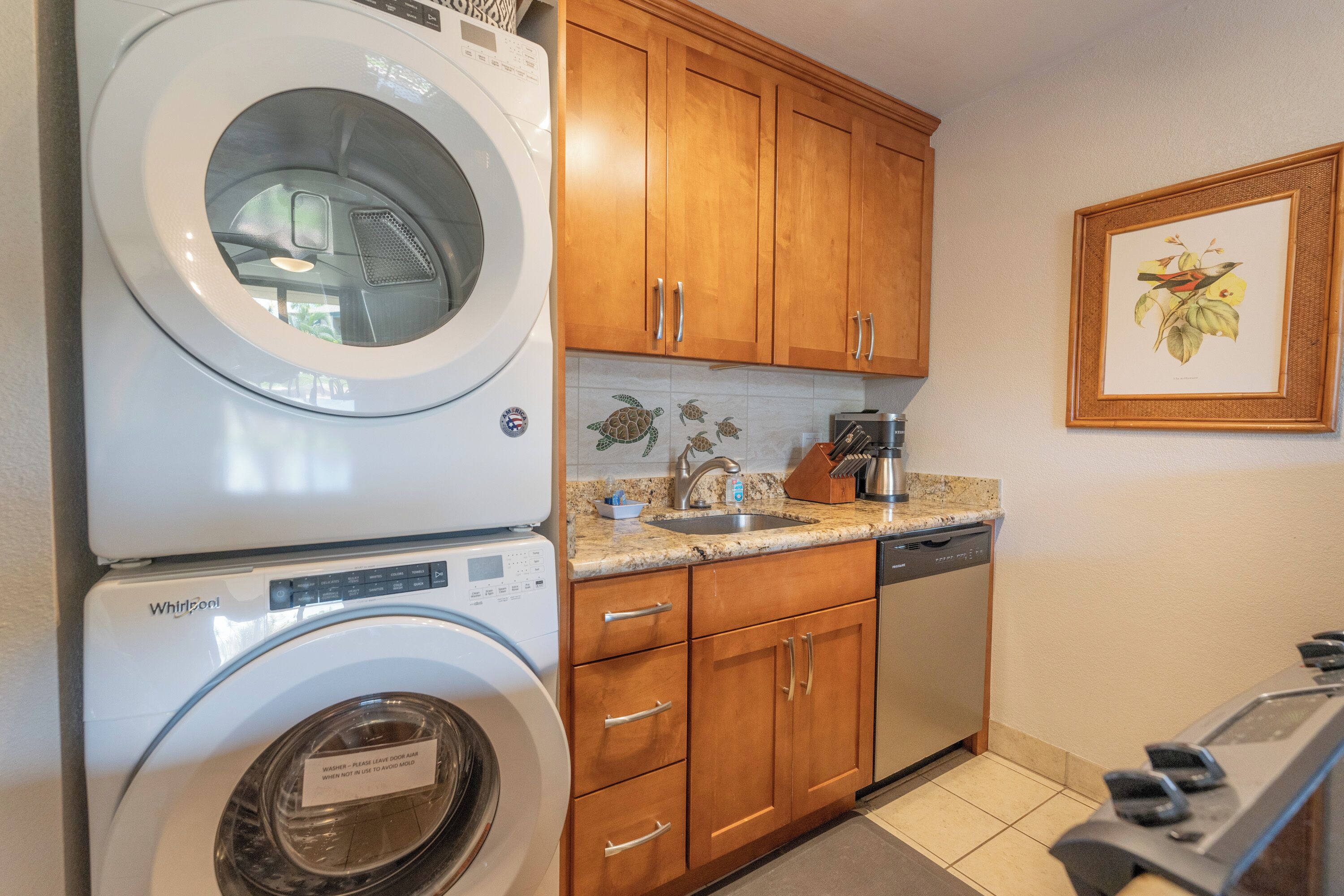 Une cuisine compacte dotée d’un lave-linge et d’un sèche-linge intégrés, de meubles en bois, d’un plan de travail en granit et d’une estampe botanique encadrée au mur.