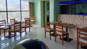 A dining area with wooden tables and chairs, a cushioned bench, and large windows offering a view of the outside. - Hotel Autumn Crocus (Guwahati)