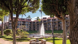 A courtyard with a fountain, surrounded by trees and a building with multiple balconies. - Vacation Retreat at Kamaole Sands, Steps from Kamaole Beach III! (Kihei)