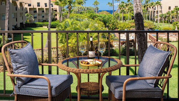 A balcony with wicker chairs, a small round table with a bottle of wine and a glass, and a view of a resort with palm trees and buildings. - Vacation Retreat at Kamaole Sands, Steps from Kamaole Beach III! (Kihei)