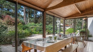 Un espace de restauration en plein air avec une table dressée pour quatre personnes, entouré de grandes fenêtres donnant sur un jardin et des arbres.