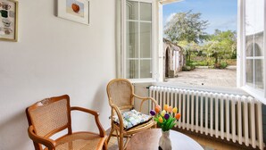 Een kamer met een rieten stoel, een tafel met een vaas tulpen en uitzicht op een tuin door een raam.