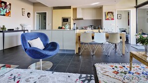 A modern kitchen with a central island, a dining area with a blue chair, and a living space with a blue armchair. - Sérénité au fil de L'eau: Appartement de Standing (L'Isle-sur-la-Sorgue)