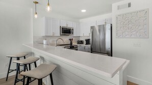 A modern kitchen with white cabinetry, a stainless steel refrigerator, a microwave, and a gas stove. There are three bar stools and a decorative wall piece. - Amazing View, Gulf View, 1Br/1Ba, Sleeps 4, Free Beach Setup + Free Tickets! (Seacrest)