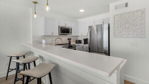 A modern kitchen with white cabinetry, a stainless steel refrigerator, a microwave, and a gas stove. There are three bar stools and a decorative wall piece. - Amazing View, Gulf View, 1Br/1Ba, Sleeps 4, Free Beach Setup + Free Tickets! (Seacrest)