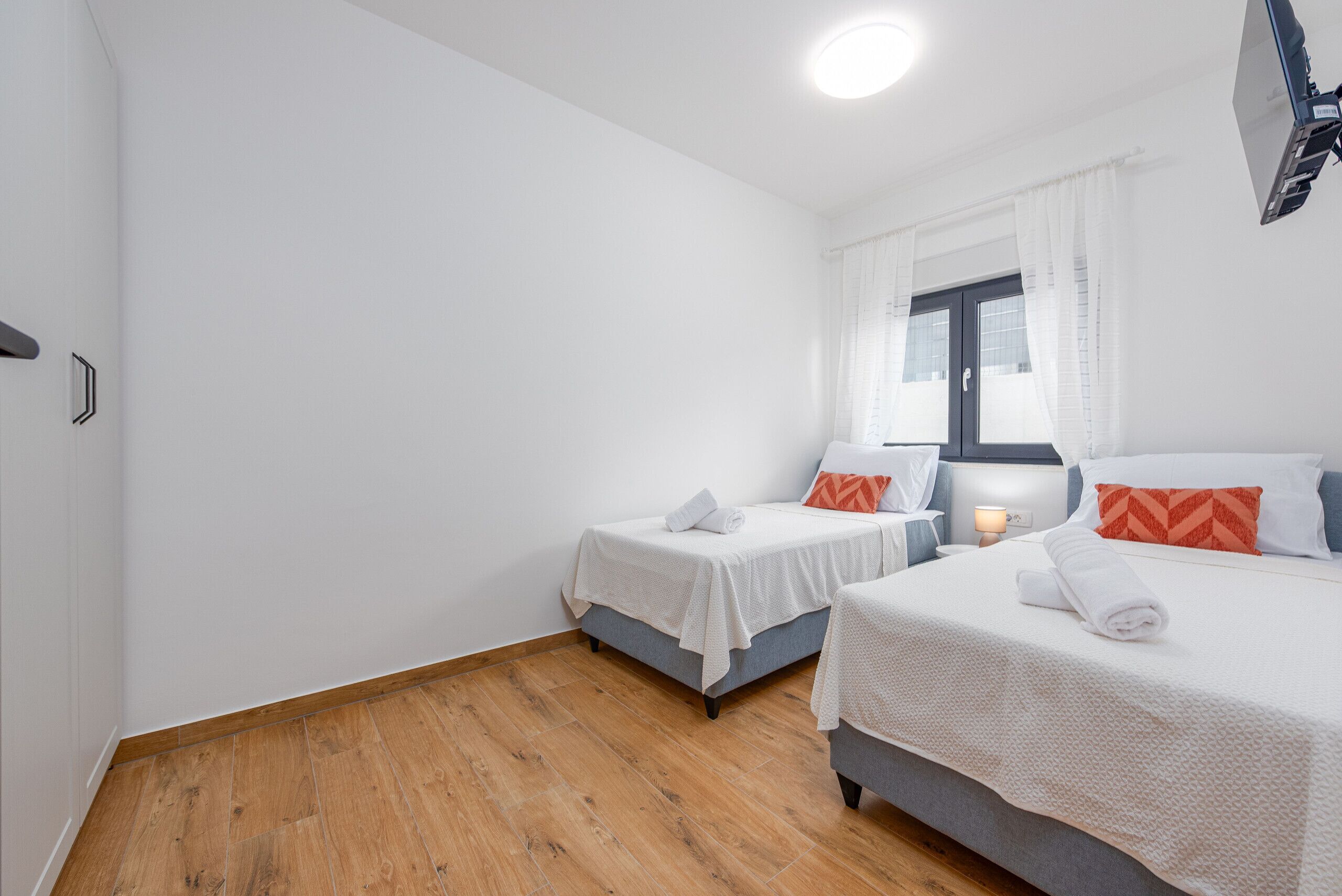 Quarto de hotel com duas camas, janela com cortinas, piso de madeira e televisão de tela plana na parede.