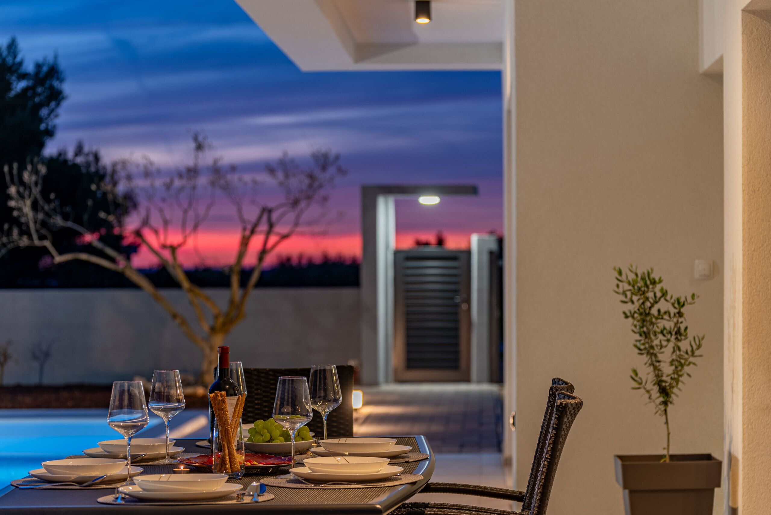 Une table dressée pour un dîner à deux, avec des verres à vin, des assiettes et une bouteille, donnant sur une piscine et un coucher de soleil.