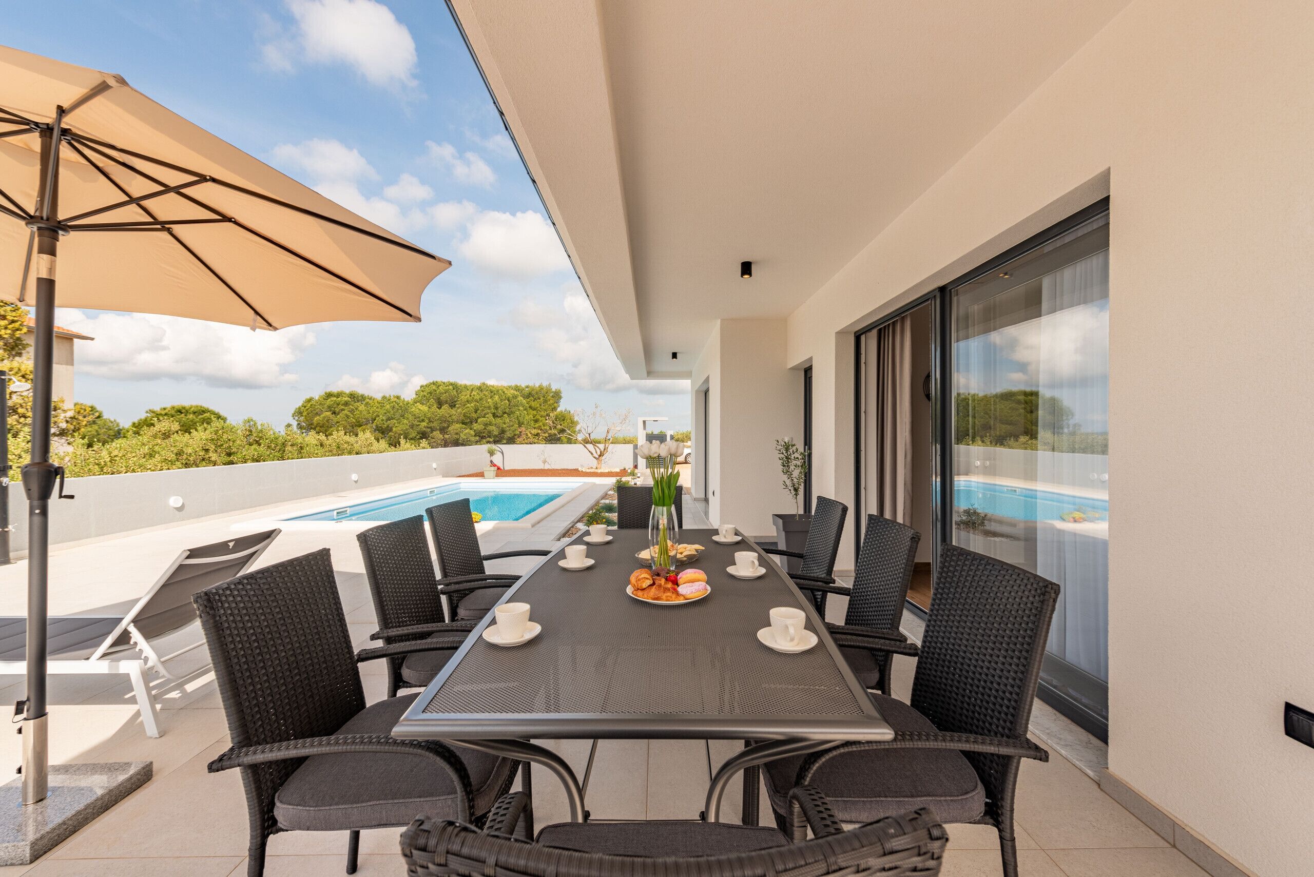 Un comedor con una mesa dispuesta para seis personas, rodeado de sillas de mimbre, un paraguas grande y vista a una piscina.