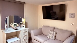 Una sala moderna con un sofá beige, un escritorio blanco pequeño con cajones, un espejo y un televisor de pantalla plana montado en la pared.