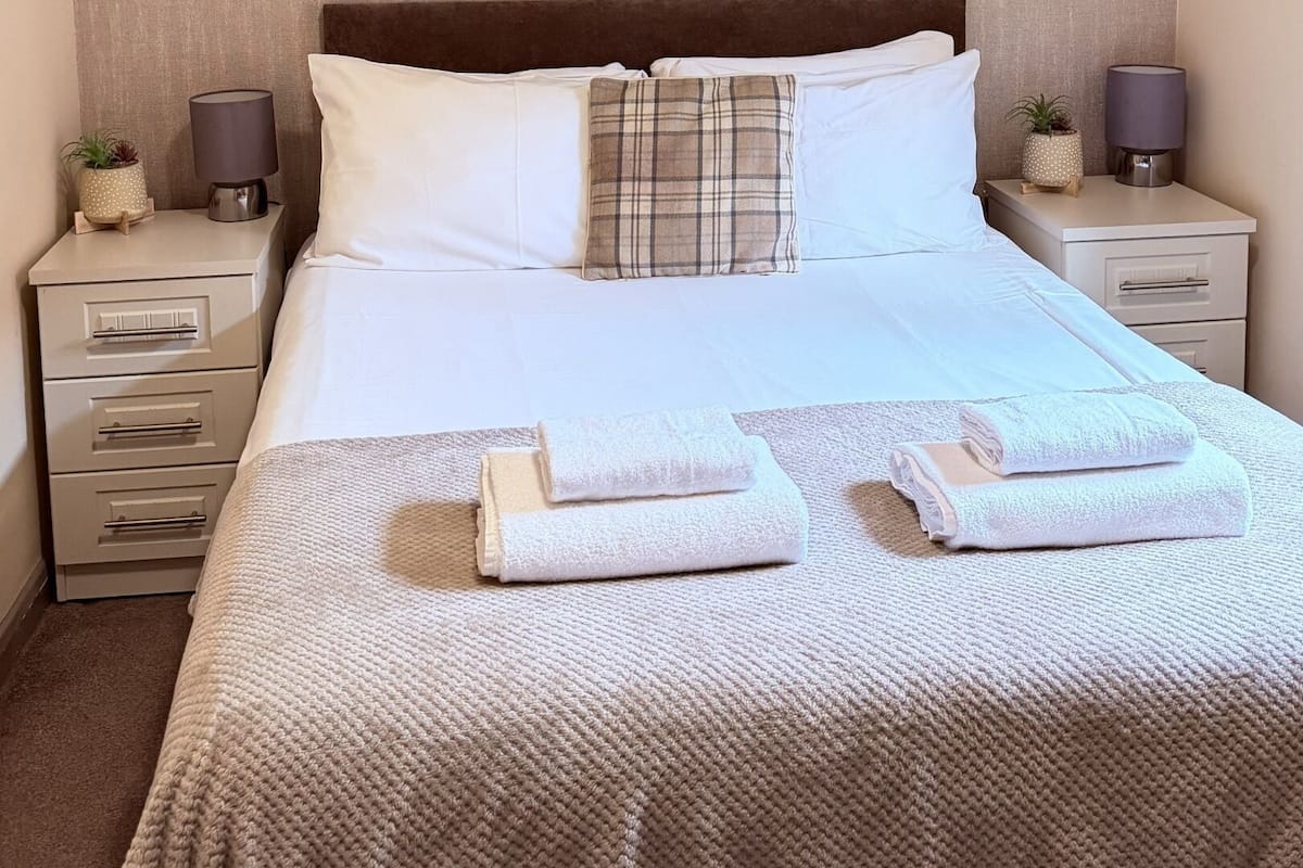 Cama bem arrumada com roupa de cama branca, um travesseiro xadrez e um mapa decorativo na parede. Há duas mesas de cabeceira com luminárias e pequenas plantas em vasos.