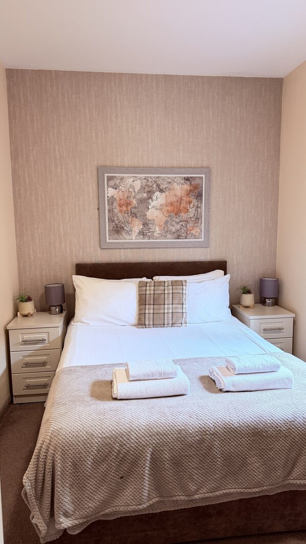 Una cama impecable con ropa de cama blanca, una almohada de cuadros y un mapa decorativo en la pared. Hay dos mesitas de noche con lámparas y pequeñas macetas.