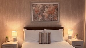 Una cama bien hecha con sábanas blancas y toallas dobladas, dos mesitas de noche con lámparas y plantas en macetas, un mapa mundial enmarcado en la pared y un cabecero de madera.
