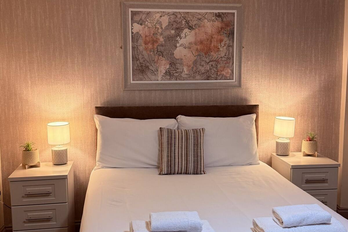 Cama bem arrumada com lençóis brancos e toalhas dobradas, duas mesas de cabeceira com abajures e plantas em vasos, um mapa-múndi emoldurado na parede e uma cabeceira de madeira.