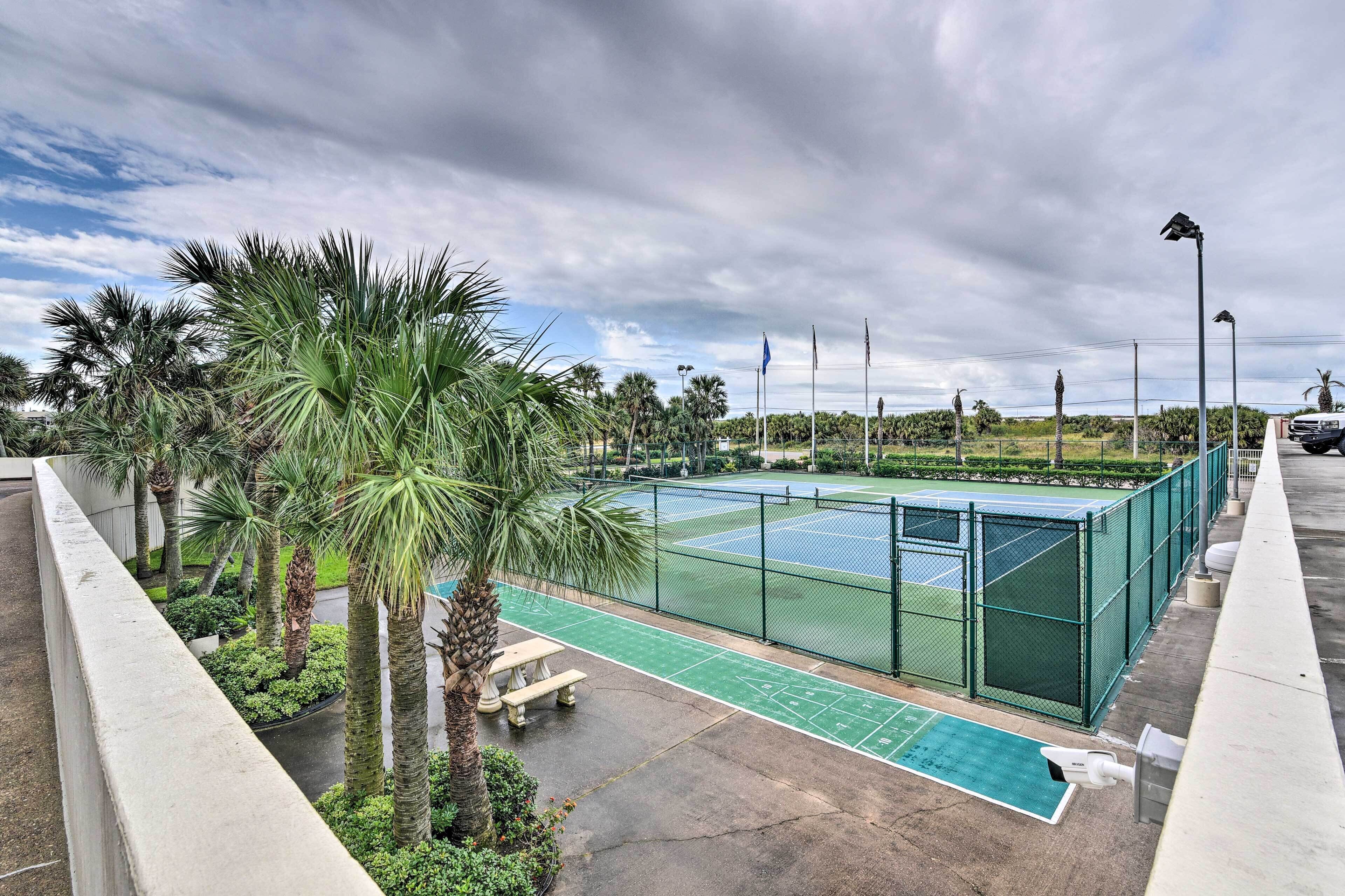 Una cancha de tenis con valla verde, palmeras y una mesa de picnic.