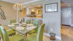 Un coin repas avec une table à plateau en verre, des chaises à revêtement vert, un lustre et une cuisine en arrière-plan.