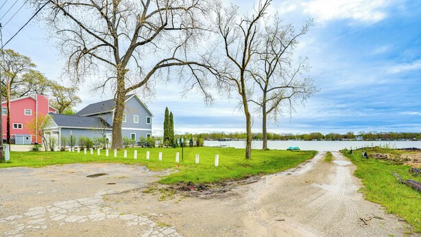 A gravel road leading to a house with a blue roof, surrounded by trees and a body of water in the background. - Kayak & Fish: 1 Block to Lakeside Park & Ramp! (Dowagiac)