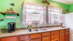 A kitchen with wooden cabinets, a coffee maker, a sink, and a window with floral curtains. - Kayak & Fish: 1 Block to Lakeside Park & Ramp! (Dowagiac)