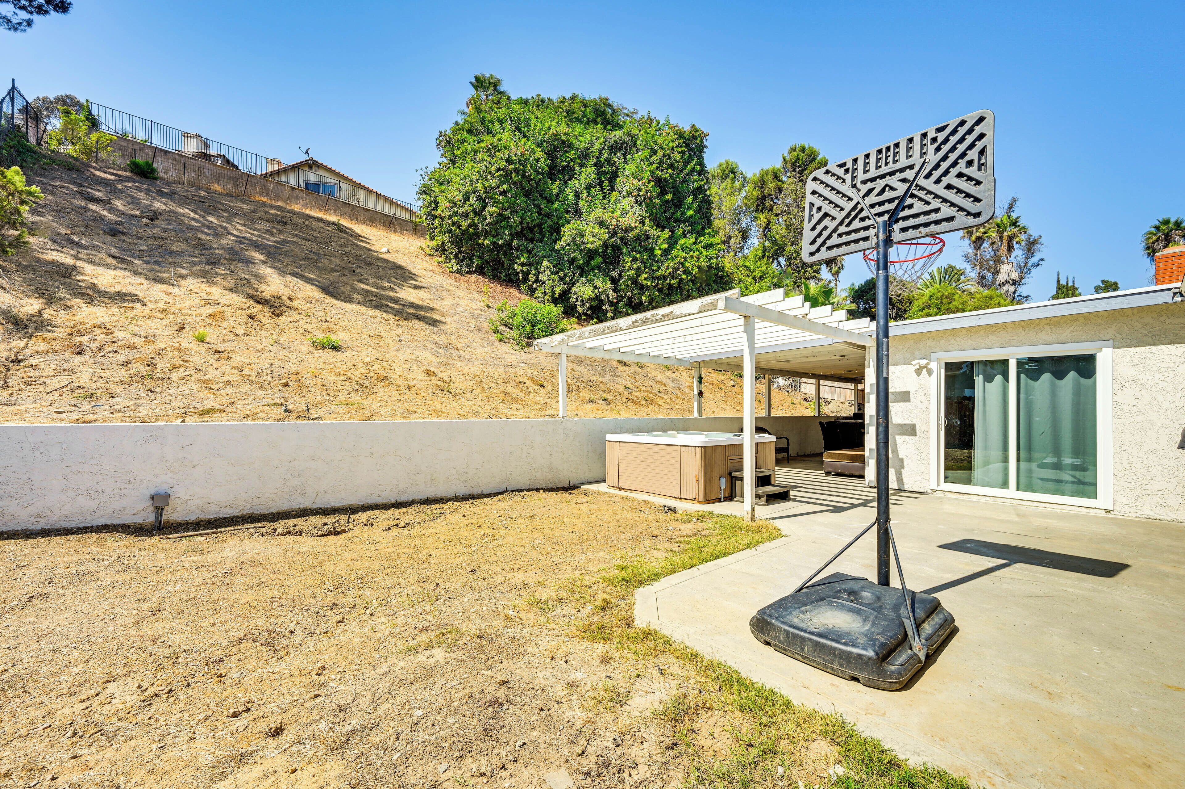 Un jardin à l’arrière d’une maison, avec un panier de basketball, un patio couvert et une colline en arrière-plan.