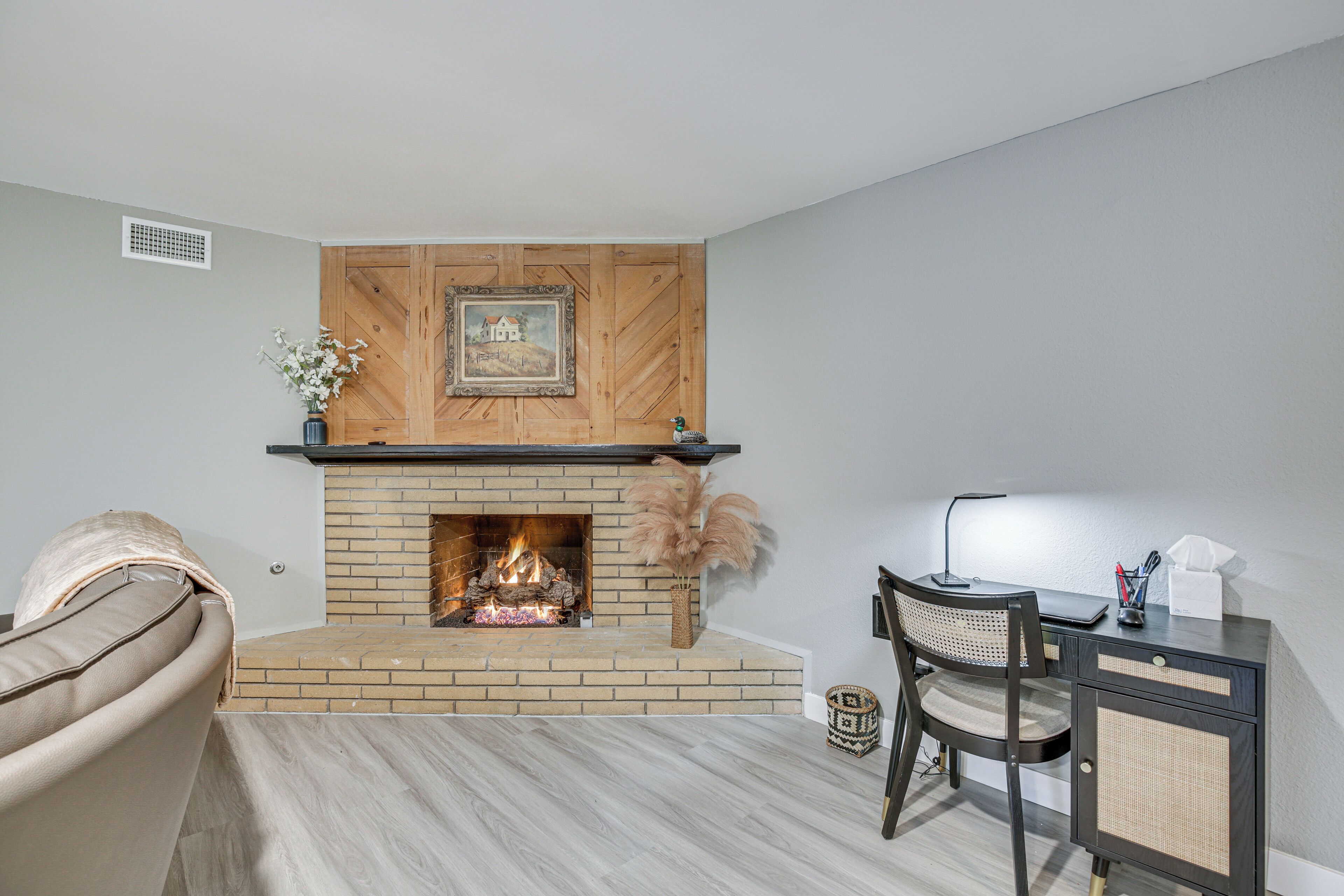 A room with a brick fireplace, a desk with a chair, and a cushioned armchair.