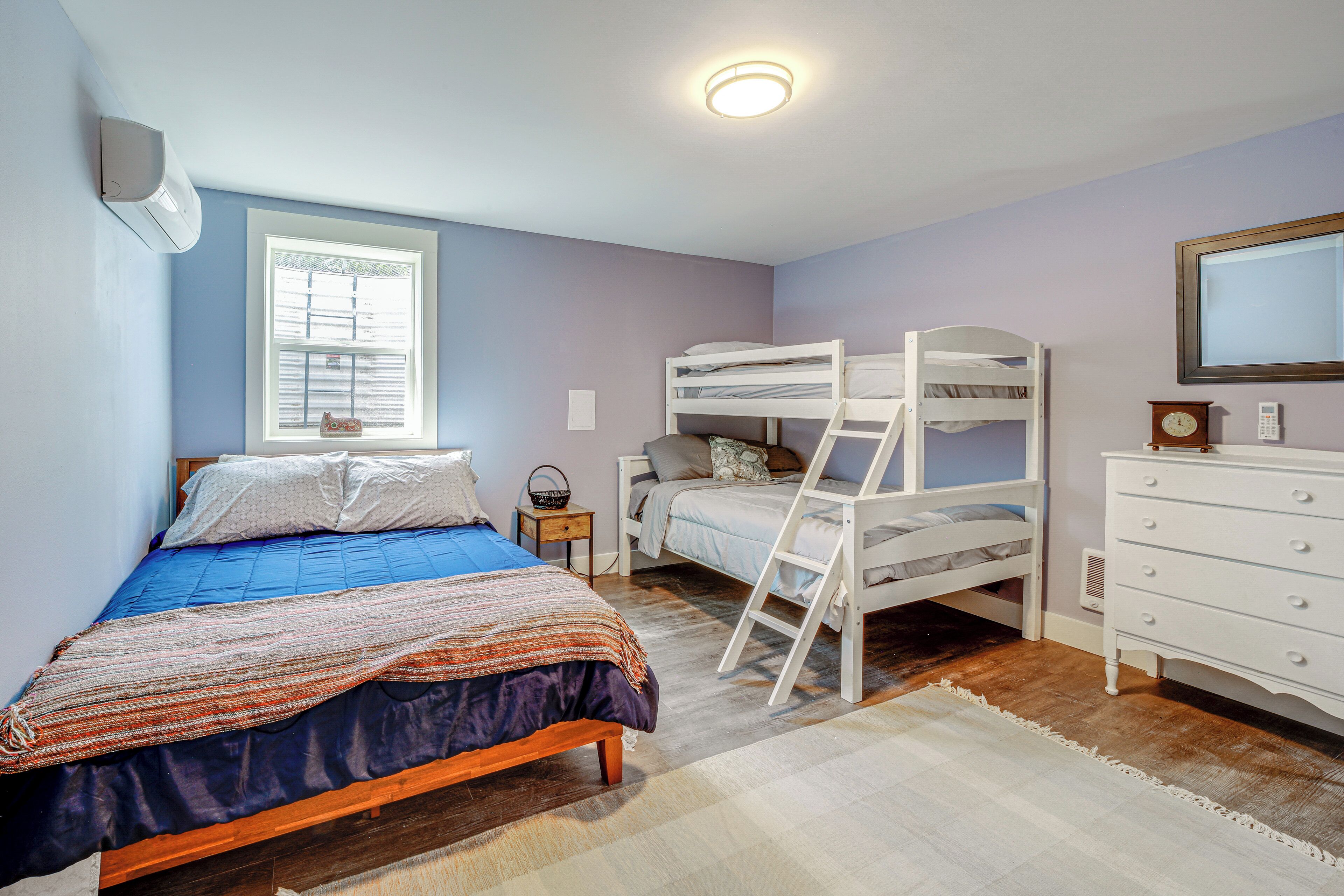 Un dormitorio con cama individual, literas, cómoda, mesita de noche, ventana con persianas y luz de techo.
